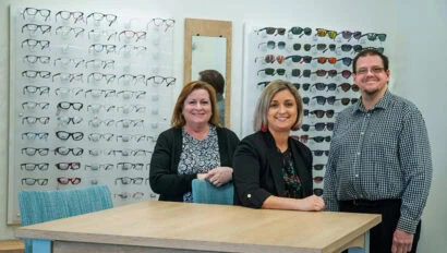 Three staff members of Simon Eye Rehoboth in front of glasses displayed on a wall.